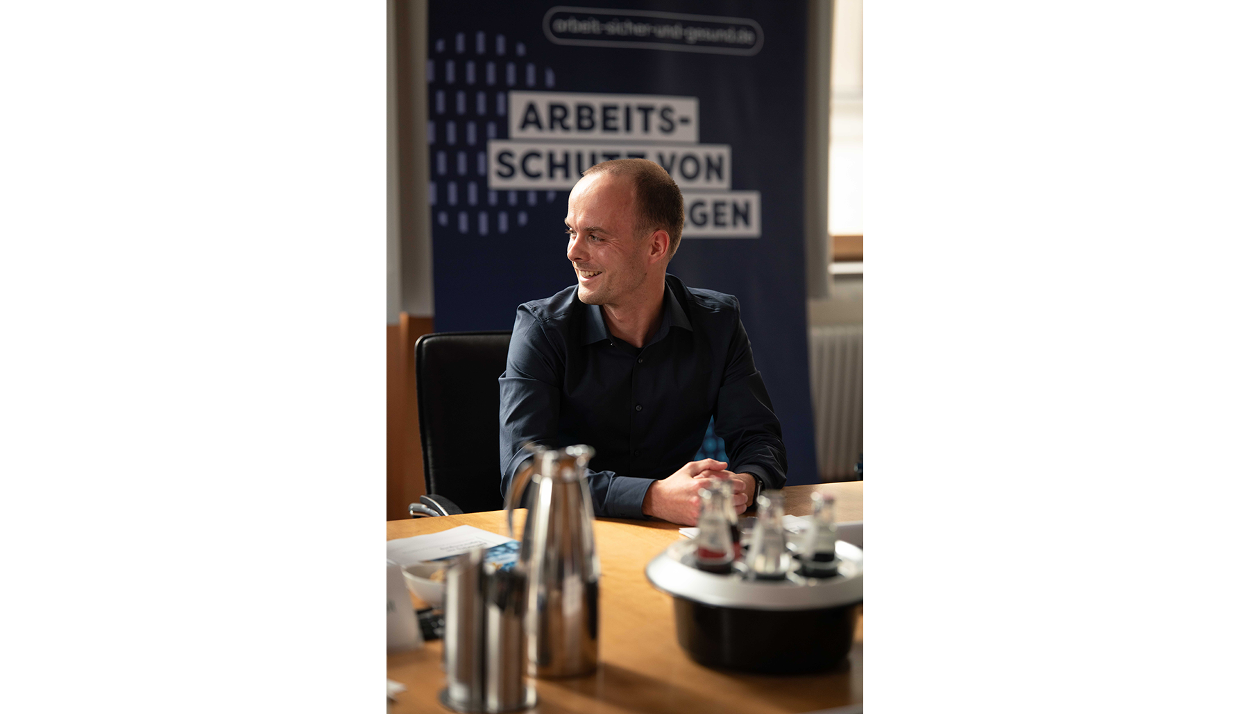 Robert Grüning sitzt am Konferenztisch. Er wendet sich lächelnd einer Person außerhalb des Bildes zu. Im Hintergrund steht ein Banner mit der Aufschrift „Arbeitsschutz von morgen“.