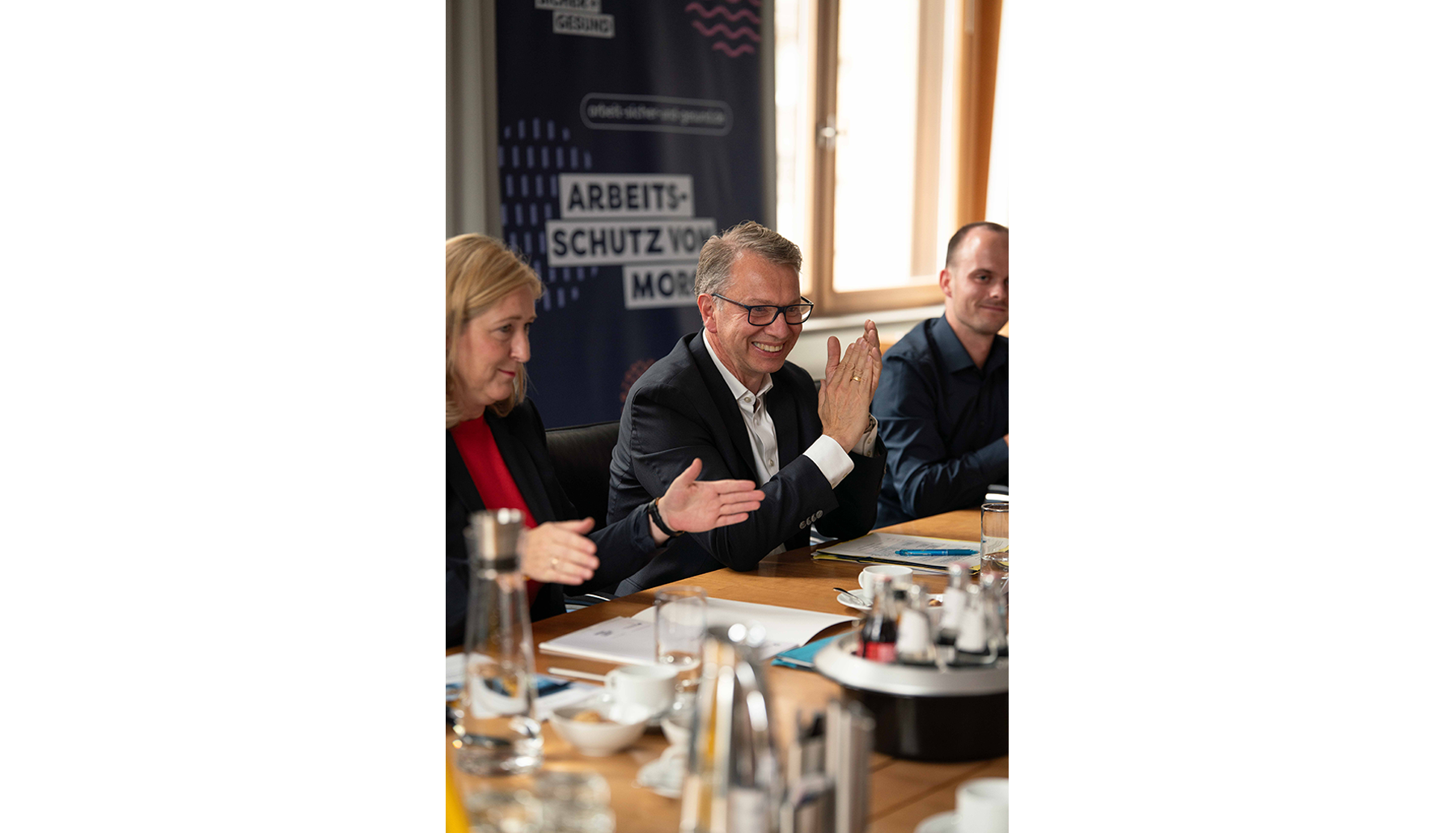 Lilian Tschan, Peer-Oliver Villwock und Robert Grüning sitzen am Konferenztisch und applaudieren. Im Hintergrund steht ein Banner mit der Aufschrift „Arbeitsschutz von morgen“.