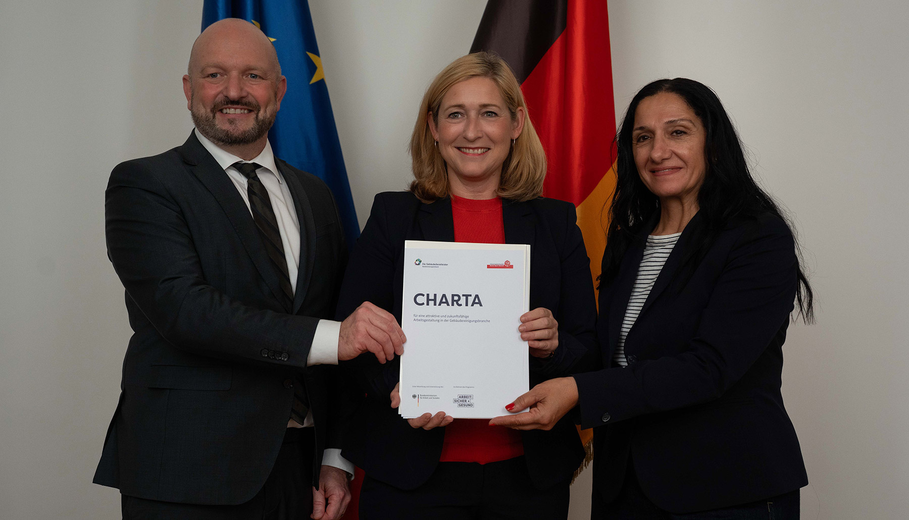 Wolfgang Molitor, Lilian Tschan und Zeynep Bicici halten gemeinsam die Charta zur Tagesreinigung hoch. Hinter ihnen stehen die Flaggen der Europäischen Union und der Bundesrepublik Deutschland. 