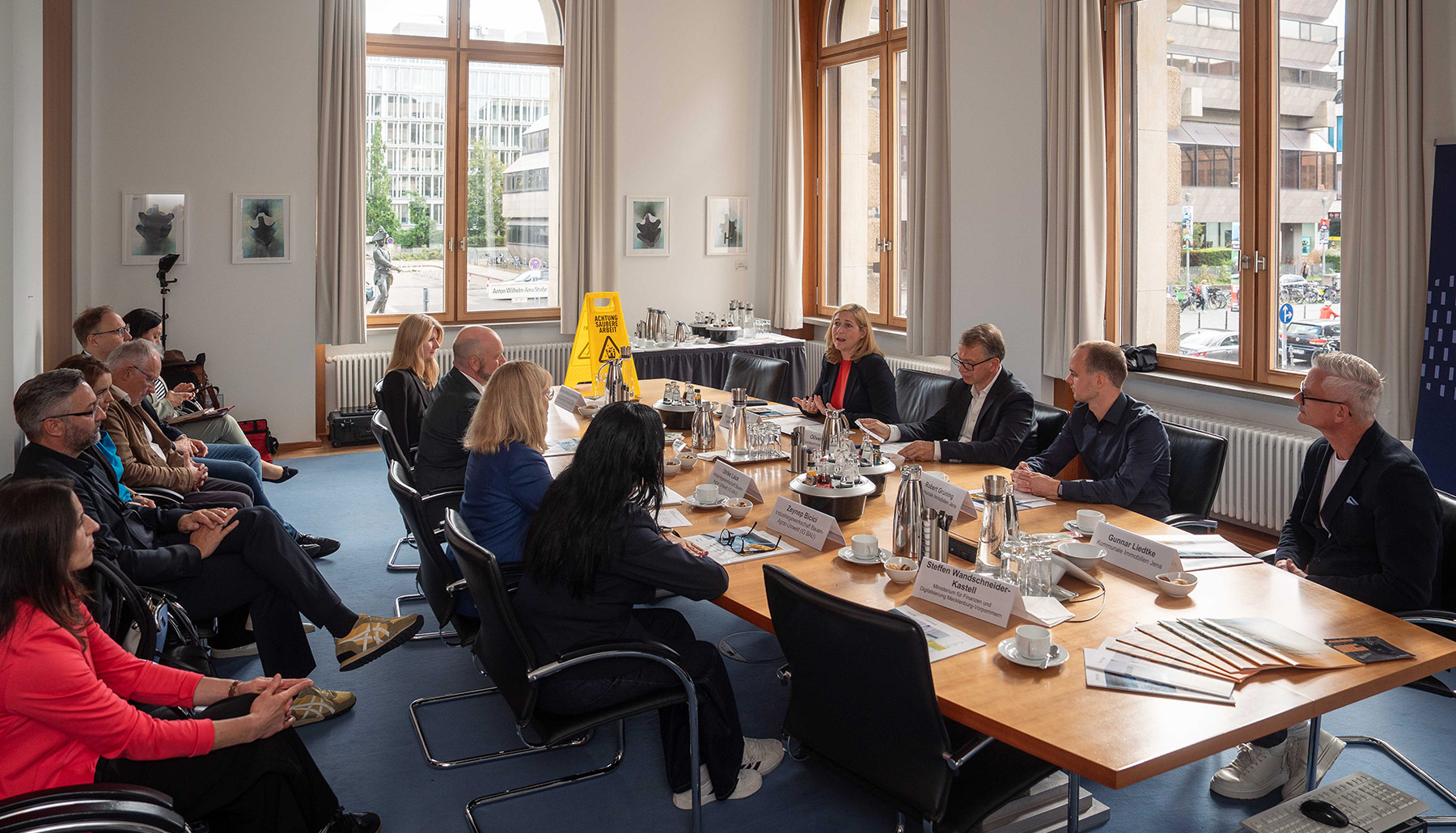 Lilian Tschan spricht zu den Teilnehmenden. Der ganze Konferenzraum und alle Teilnehmenden sind sichtbar. Die Erstunterzeichnenden sitzen am Tisch, weitere Teilnehmende in einer zweiten Reihe.