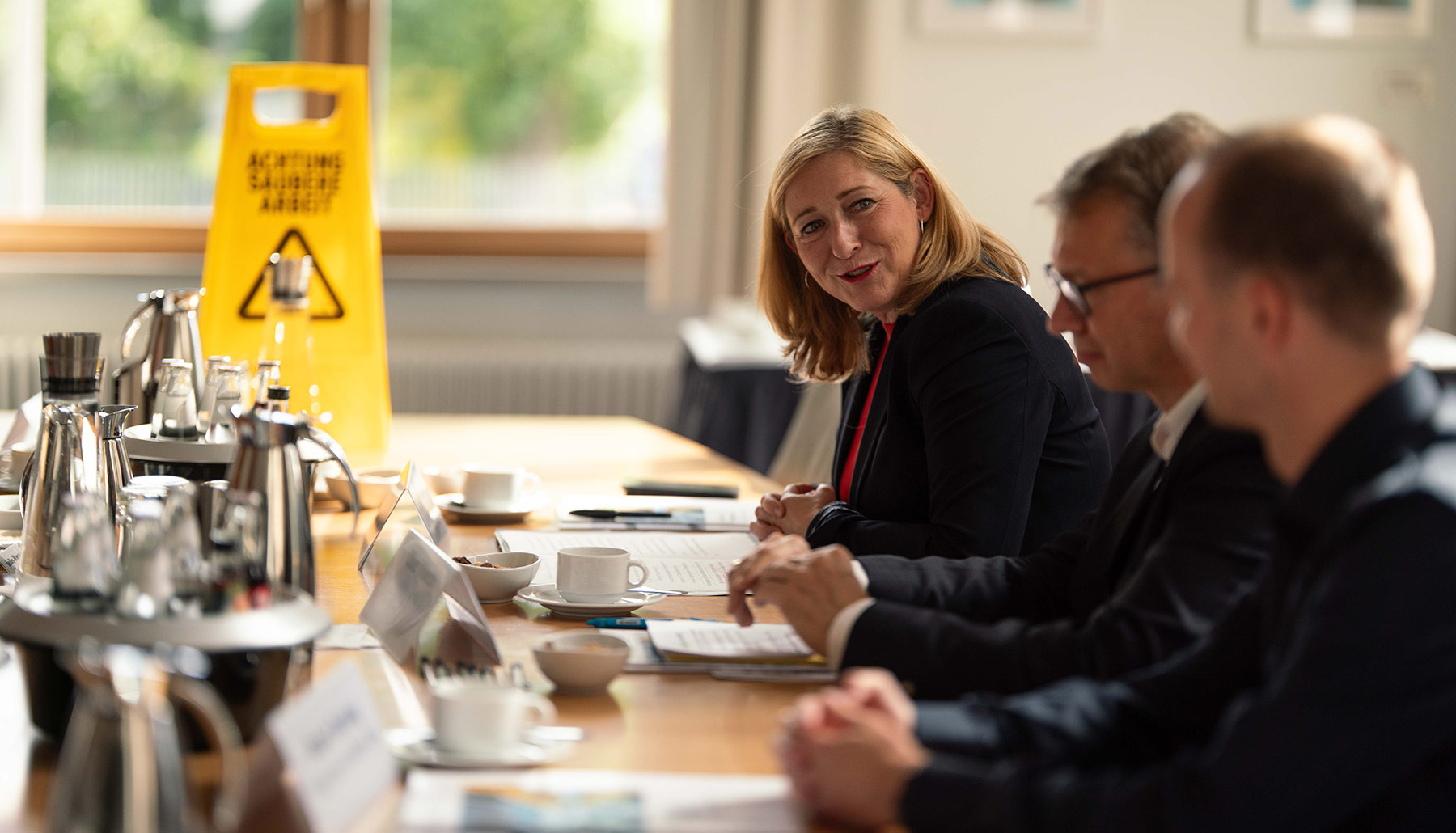 Lilian Tschan spricht zu mehreren Teilnehmenden am Konferenztisch. Sie ist fokussiert, weitere Teilnehmende unscharf. Auf dem Tisch stehen Kaffeetassen, Kannen und Wasserflaschen. Vor den Teilnehmenden liegen Dokumente.
