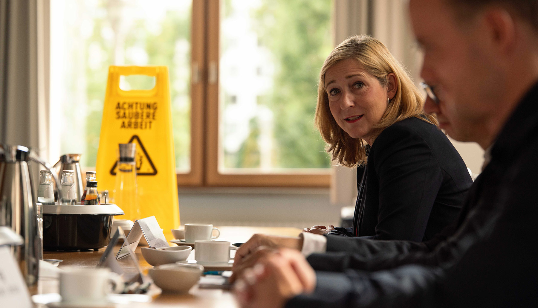 Lilian Tschan spricht zu den Teilnehmenden am Konferenztisch. Sie ist fokussiert. Im Hintergrund steht ein Aufsteller mit der Aufschrift ACHTUNG SAUBERE ARBEIT.