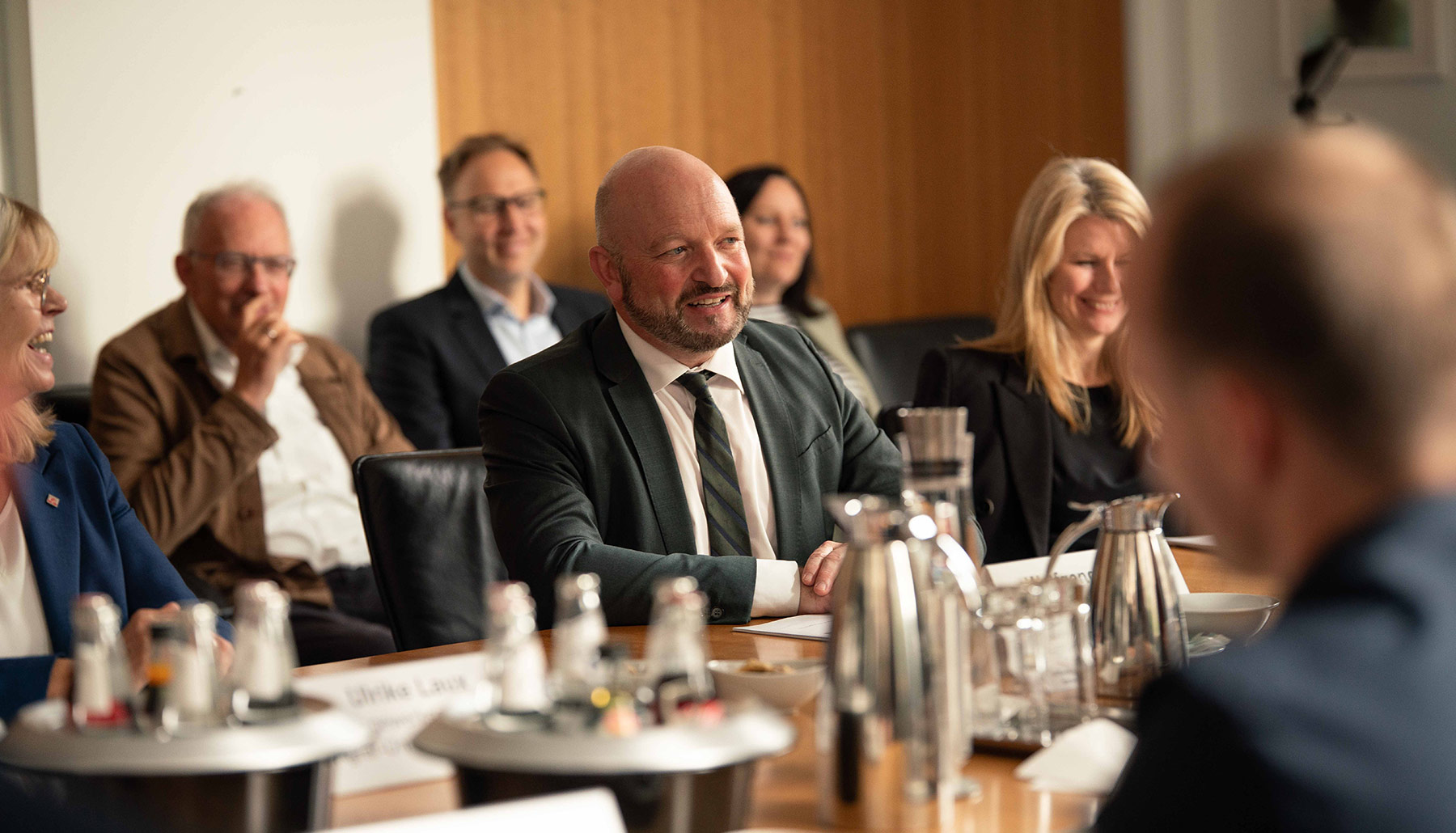 Wolfgang Molitor spricht am Konferenztisch. Er lächelt. Neben ihm und im Hintergrund sitzen weitere Teilnehmende und lachen. 