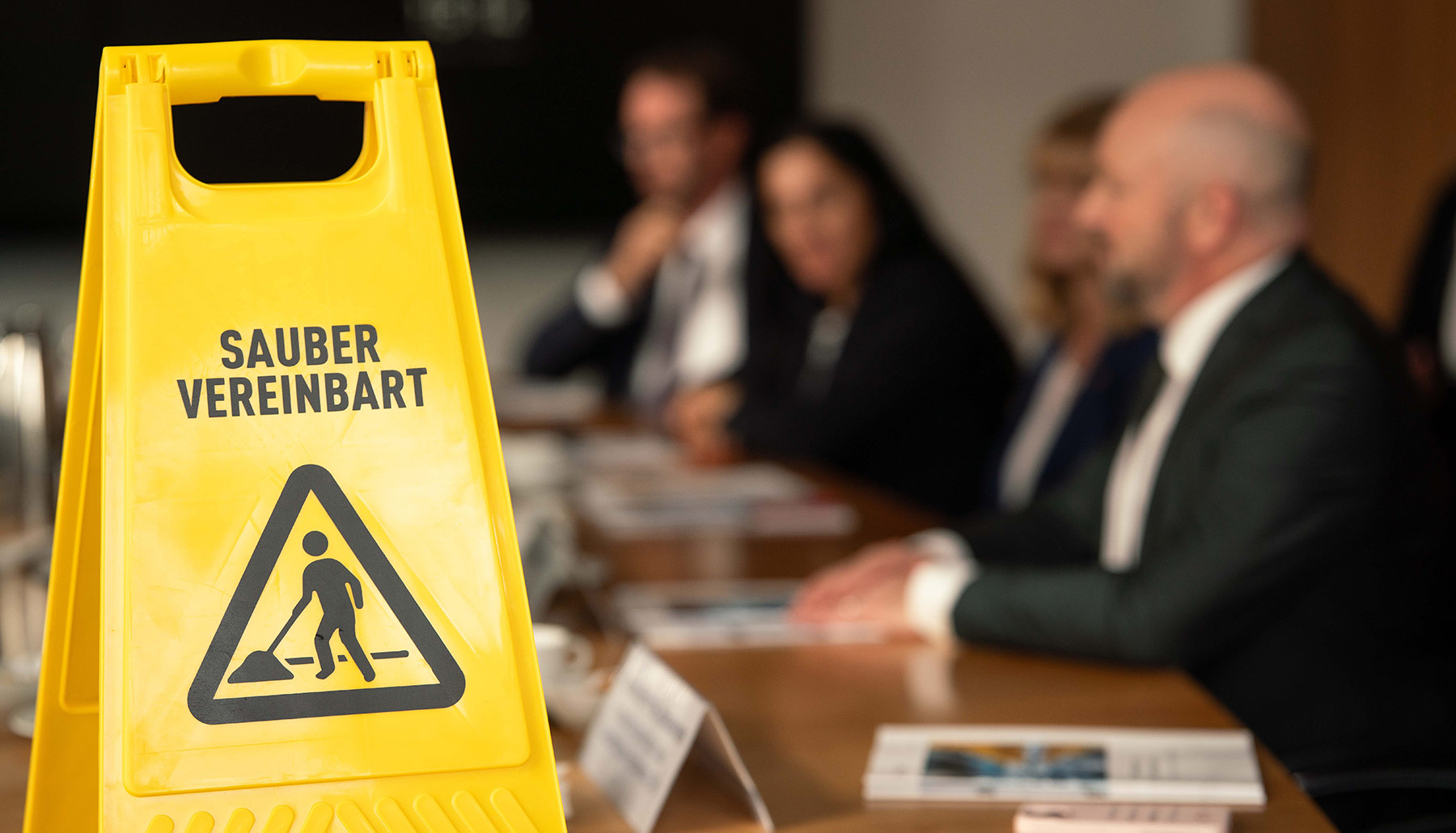 Auf dem Konferenztisch steht ein Aufsteller in Form eines Warnschilds mit der Aufschrift „Sauber vereinbart“ und einem Symbol für Reinigungsarbeit. 
