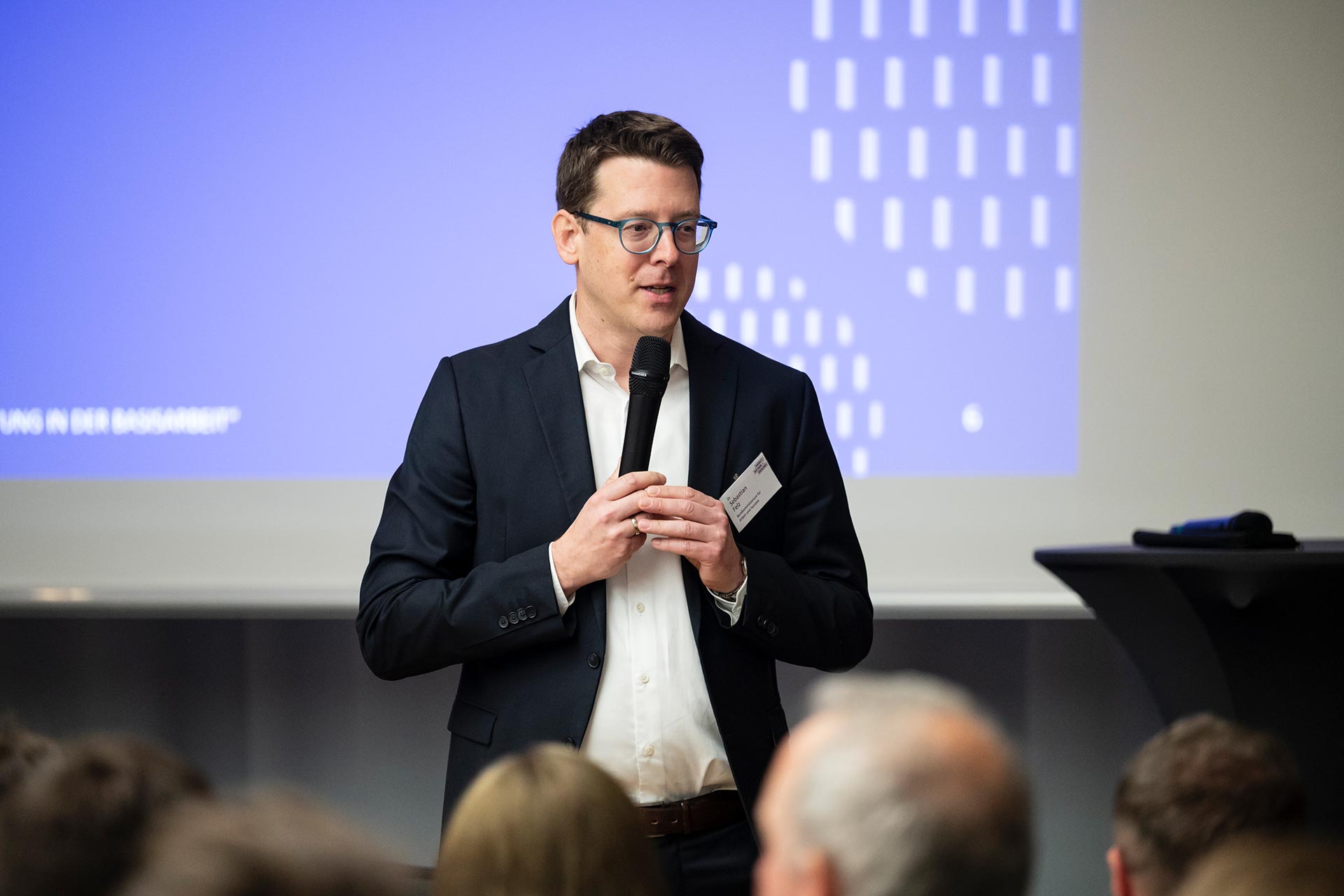 Ein Mann steht vor einer Leinwand, die eine Präsentation zeigt, und spricht in ein Mikrofon.