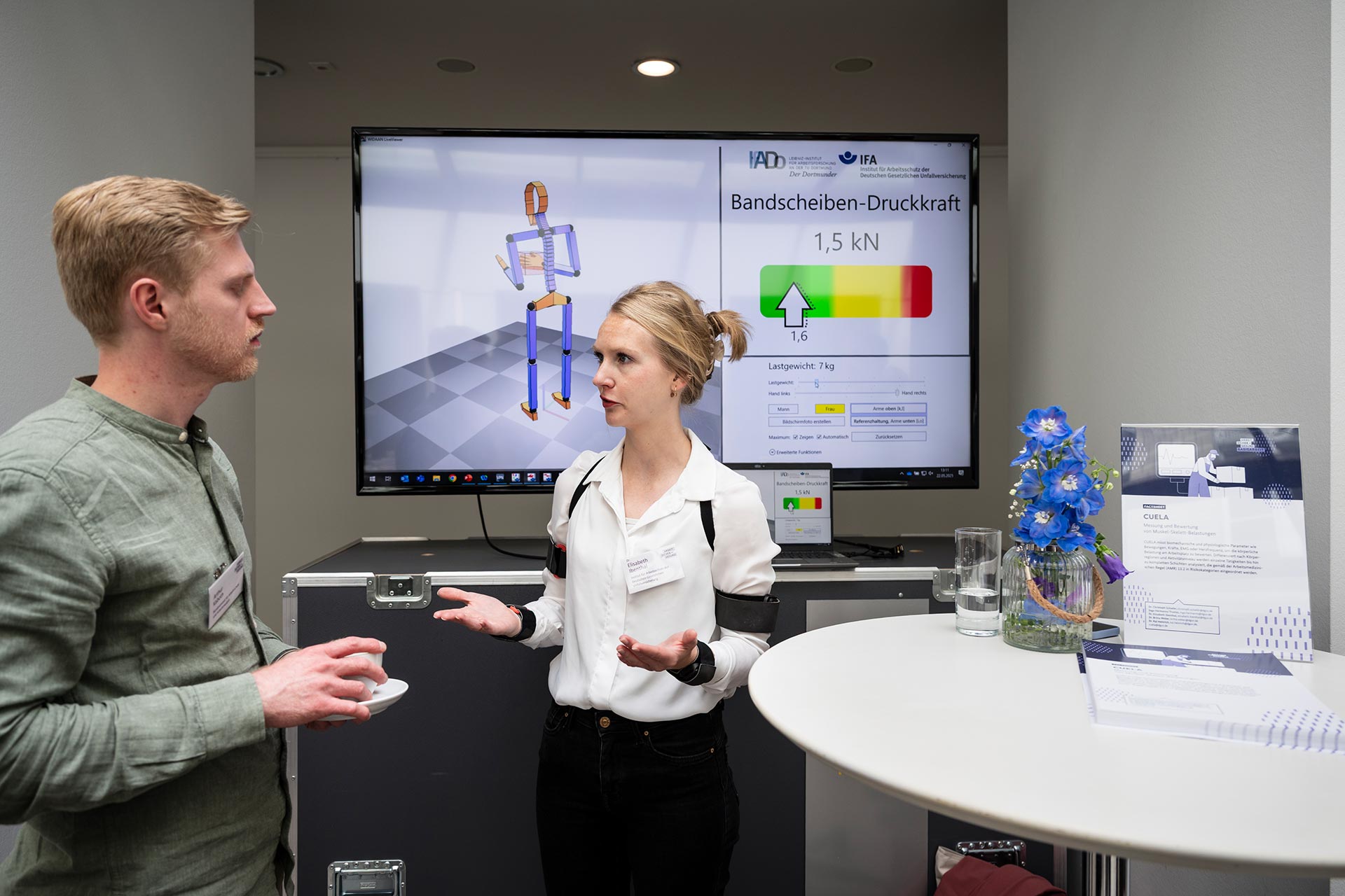 Eine Frau und ein Mann stehen vor einem großen Bildschirm und unterhalten sich . Der Bildschirm zeigt das Computermodell eines Menschen sowie eine Grafik mit dem Titel "Bandscheiben-Druckkraft".