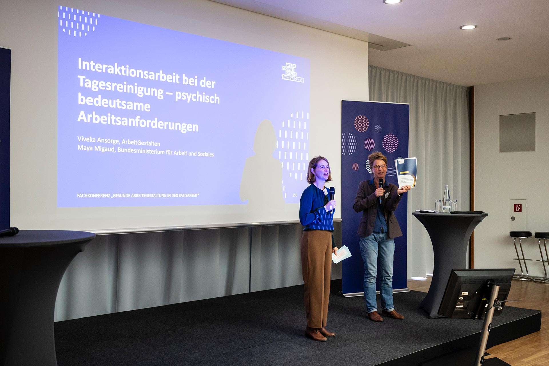Zwei Frauen sprechen in ein Mikrofon, eine von Ihnen zeigt ein bedrucktes Blatt Papier. Sie stehen vor einer Leinwand, die eine Präsentation mit dem Titel "Interaktionsarbeit bei der Tagesreinigung - psychisch bedeutsame Arbeitsanforderungen" zeigt.